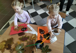 Dzieci przy stoliku lepią liście do kartek i przyczepiają oczka.