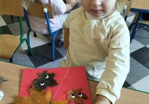 Dzieci przy stoliku lepią liście do kartek i przyczepiają oczka.