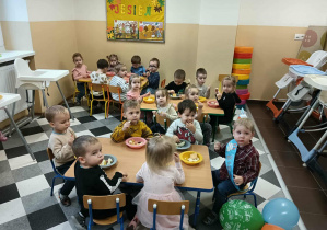 Dzieci siedzą przy stolikach i spożywają poczęstunek od jubilata.