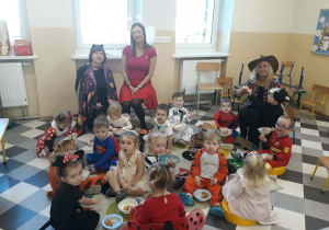 Dzieci i ciocie w przebraniach siedzą przy stolikach i spożywają posiłek.